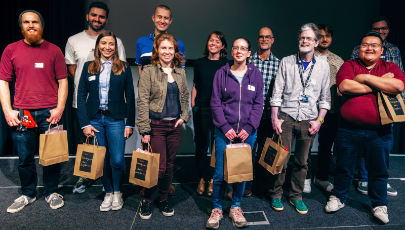 Line up of speakers from Shiny in Production 2023, all look happy and are holding brown gift bags. From left to right: Back row - Chris Brownlie, Colin Gillespie, Cara Thompson, Andrie de Vries, Liam Kalita, George Stagg; Front row - Janion Nevill, Anna Skrzydło, Clareece Nevill, Naomi Bradbury, Russ Hyde, Tan Ho.